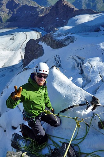 Angeseilter Hochtourengeher mit Hochtourenausrüstung lacht in die Kamera und macht ein Piece-Zeichen.