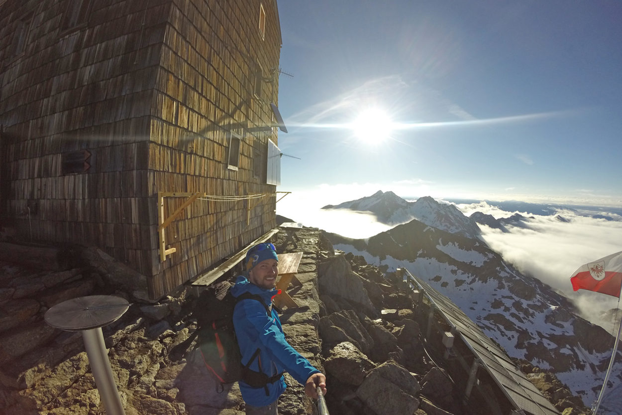 Ein Bergsteiger in der Sonne vor dem Becherhaus, im Hintergrund schneebedeckte Berge.